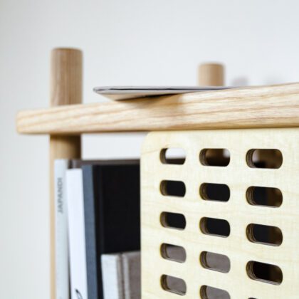 Detail eines Regals im Skandi-Stil aus Esche Massivholz mit Holzblenden mit Gitterstruktur inszeniert vor weißer Wand mit Büchern und Zeitung