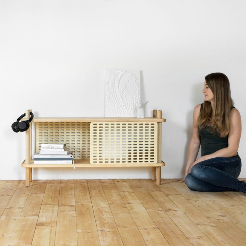 Eine Frau sitzt in einem Raum auf dem Holzboden und Blickt auf ein Lowboard aus Esche Massivholz mit Schiebeblenden aus Holz inszeniert vor weißer Wand mit Büchern, einem Gemälde, einem weißen Porzellanpinguin von halblang und Kopfhörern darauf.