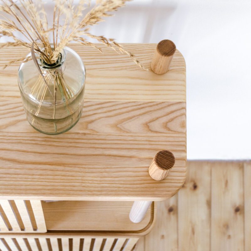 Detailsicht einer natürlichen Szene auf ein Regal aus Esche Massivholz mit Maserung und Schiebeblenden aus Holz inszeniert vor weißer Wand mit einer Vase mit Trockenblumen