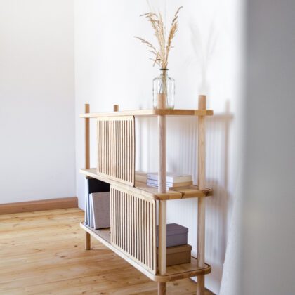 Blick in einen skandinavisch eingerichteten Raum auf ein Regal aus Esche Massivholz mit Schiebeblenden aus Holz inszeniert vor weißer Wand mit Büchern und Vase mit Trockenblumen